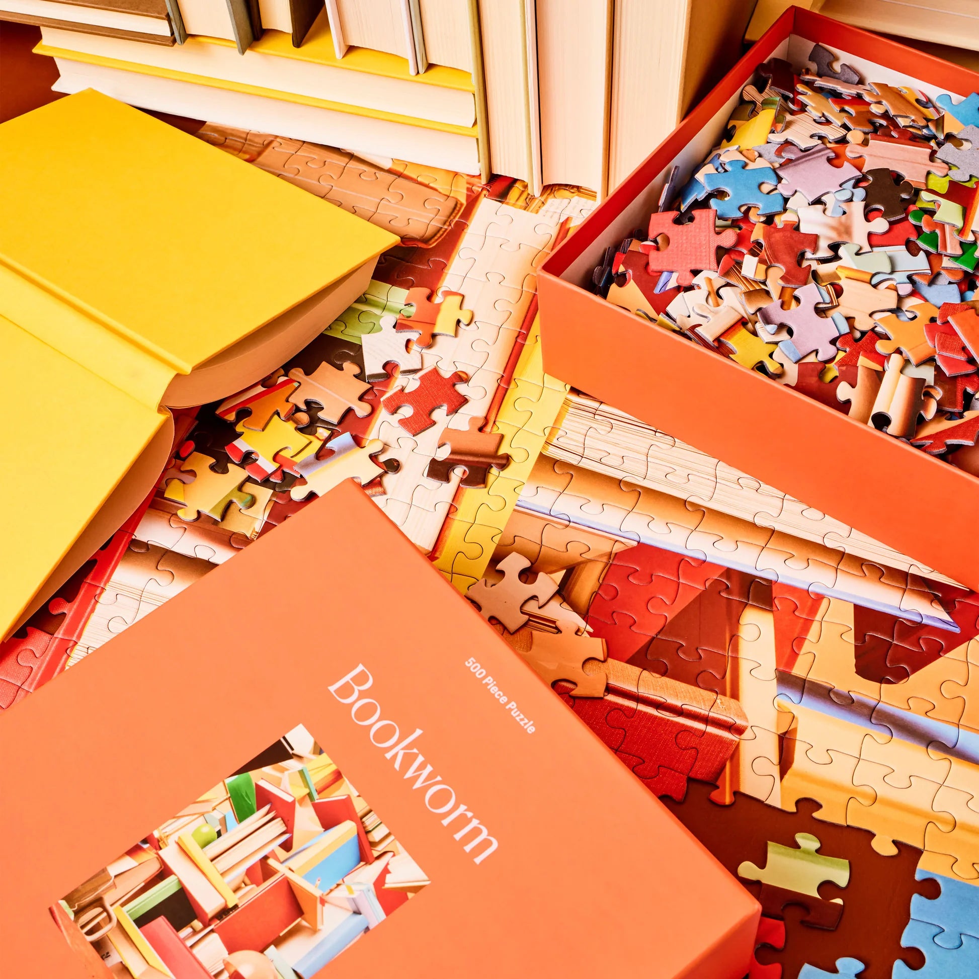 Bookworm Puzzle by Piecework Puzzles, shown open in box on floor with the puzzle put together beneath, with books in background