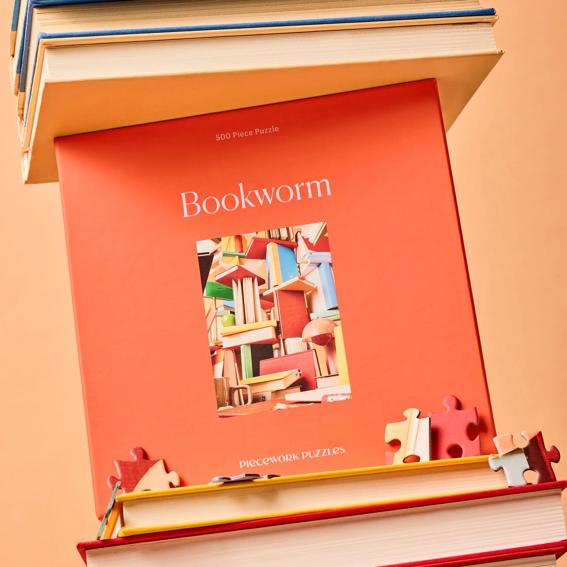 Bookworm Puzzle by Piecework Puzzles, shown in box, stacked with books, on orange yellow background