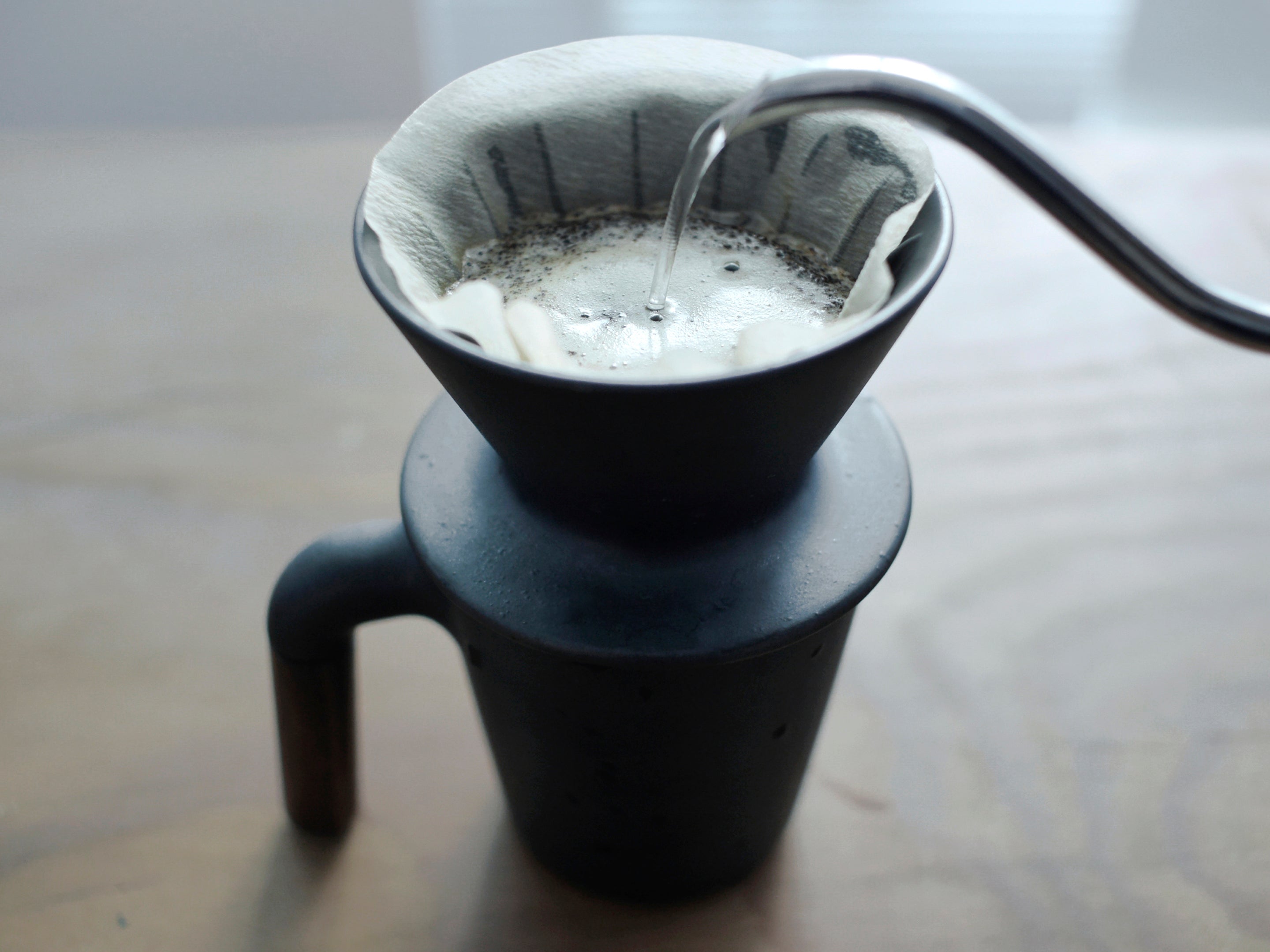Patio Ceramic Coffee Dripper by HMM in matte black, shown with coffee and filter and hot water being poured in