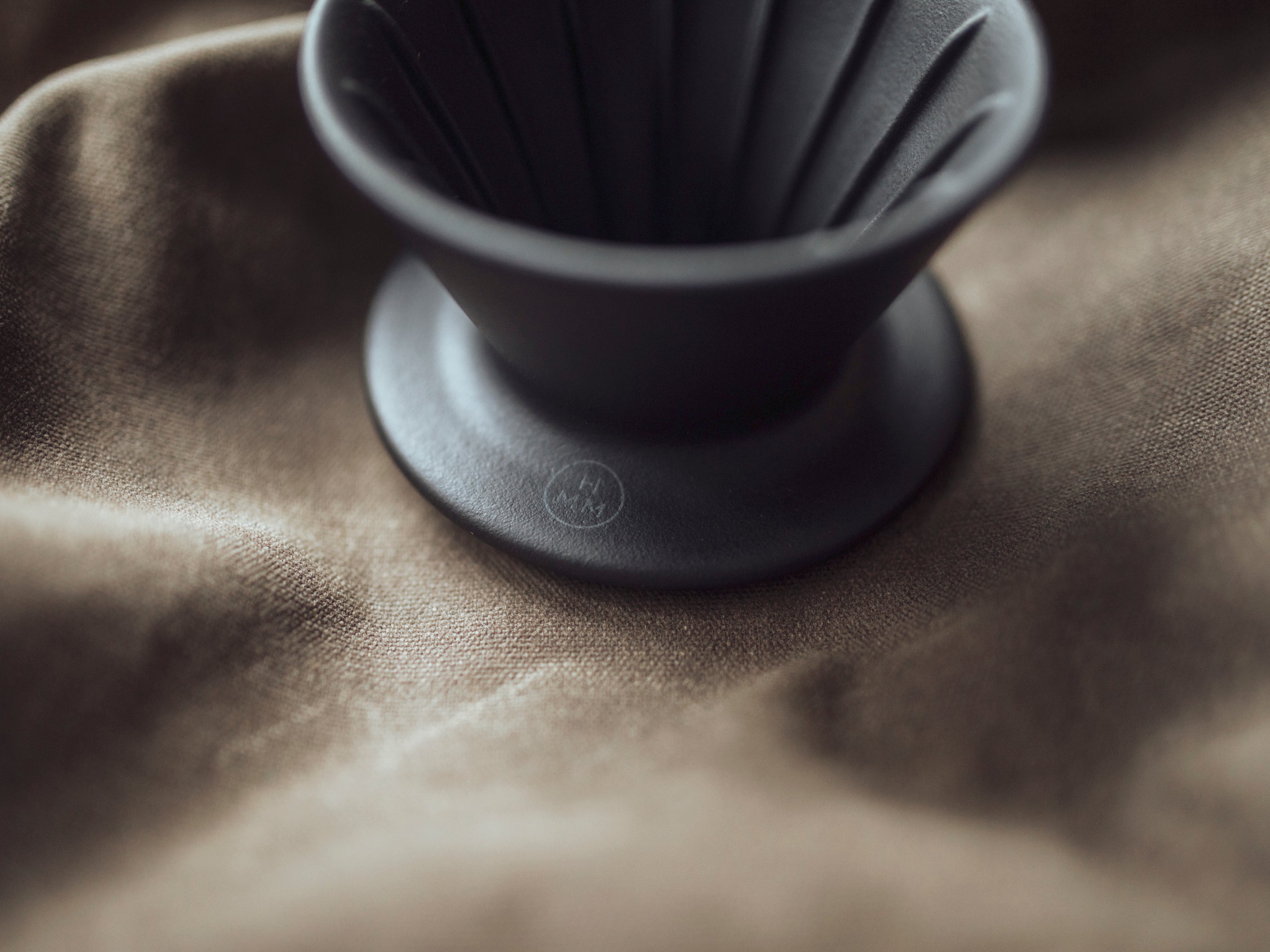 Patio Ceramic Coffee Dripper by HMM in matte black, shown from above angle, sitting on fabric, with HM logo visible