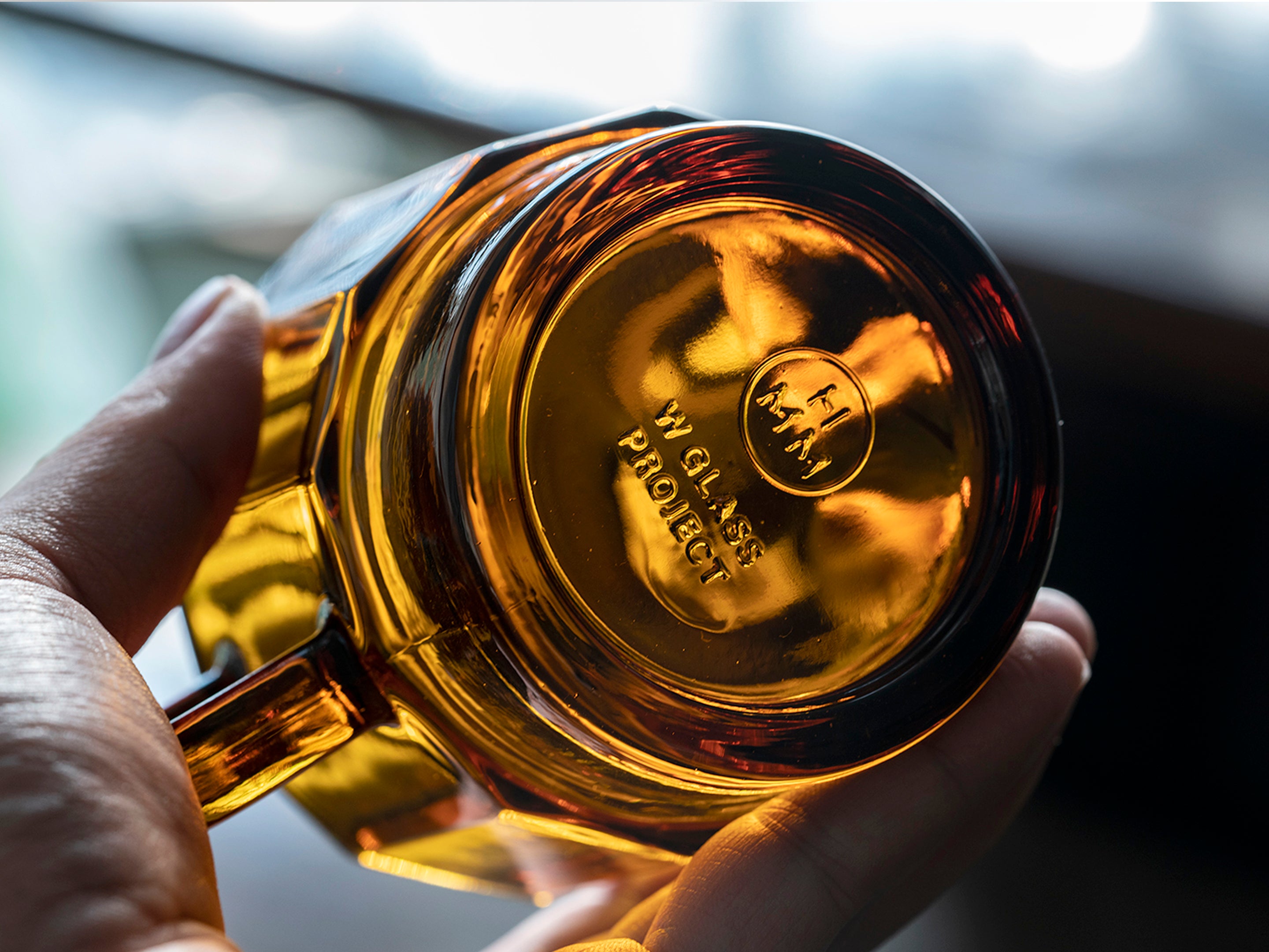 Bottom of W Glass by HMM shown in amber color, with logo visible, held up by a person's hand