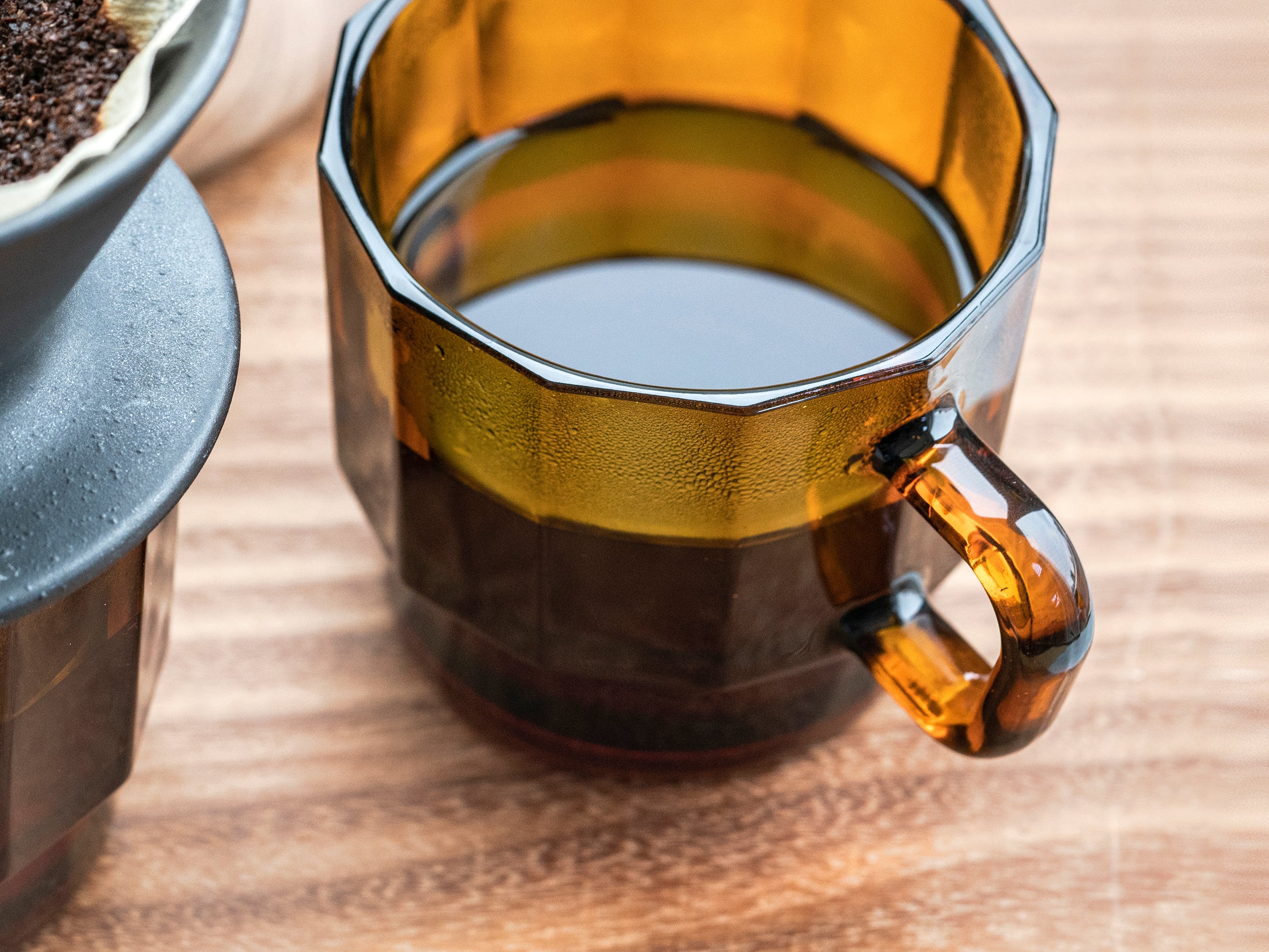 W Glass by HMM shown in amber color filled with coffee, and resting on wood surface, with coffee dripper partially in view