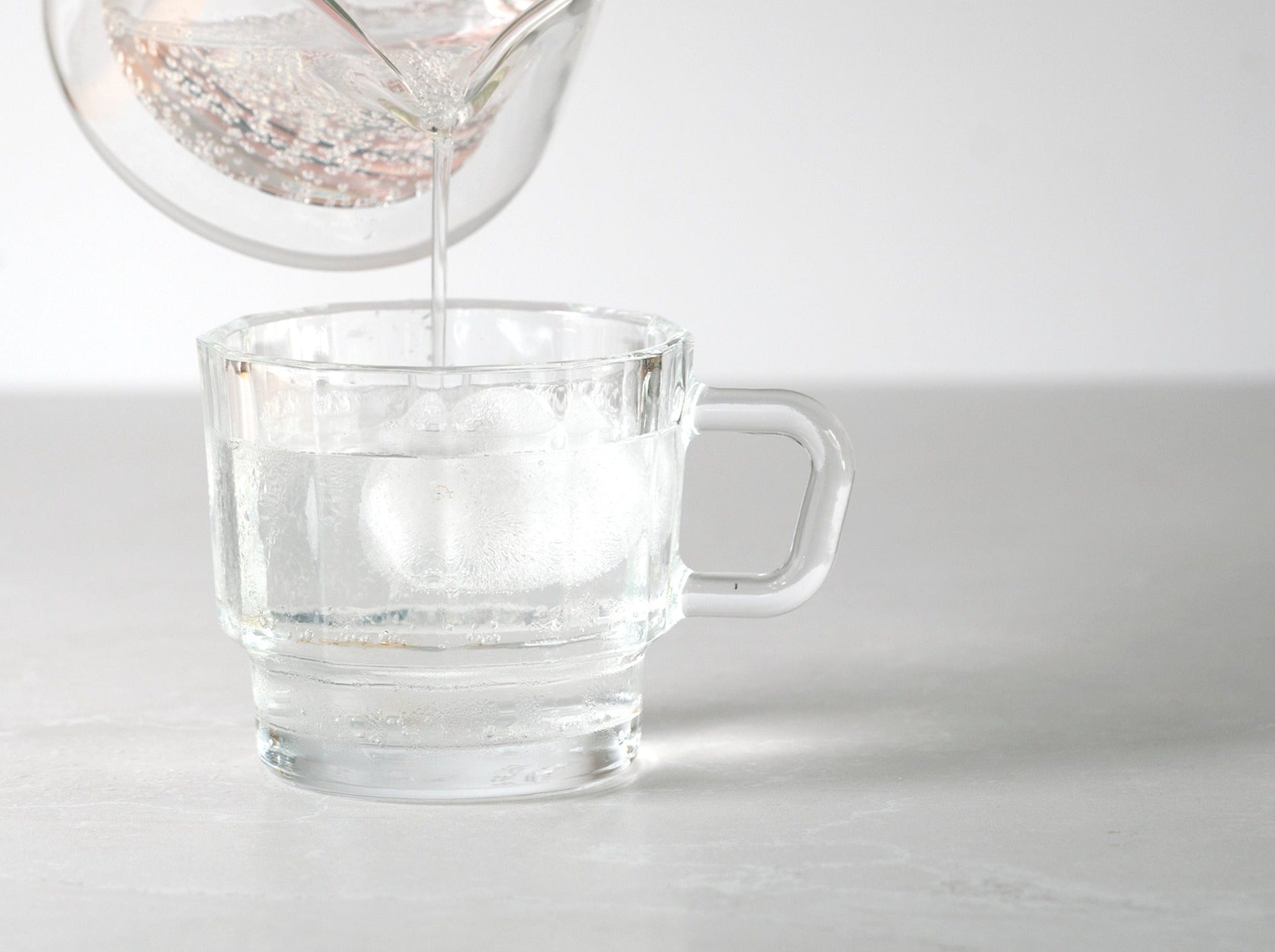 W Glass by HMM shown in clear, from side profile with ice water being poured in, on light gray background