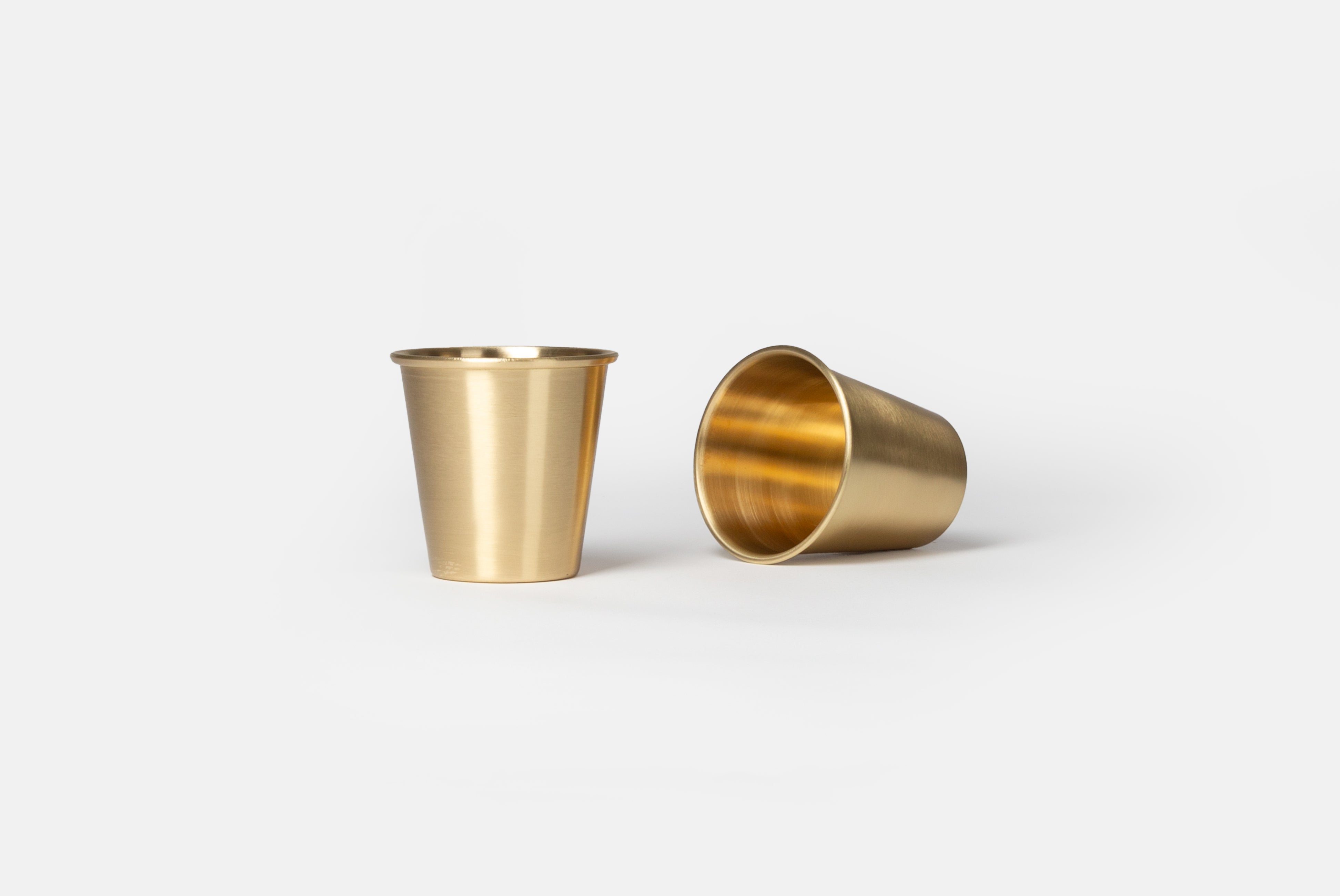 Brass Cup by Japanese Brand Fog Linen, shown as a pair with one cup standing and one laying down, on a white background