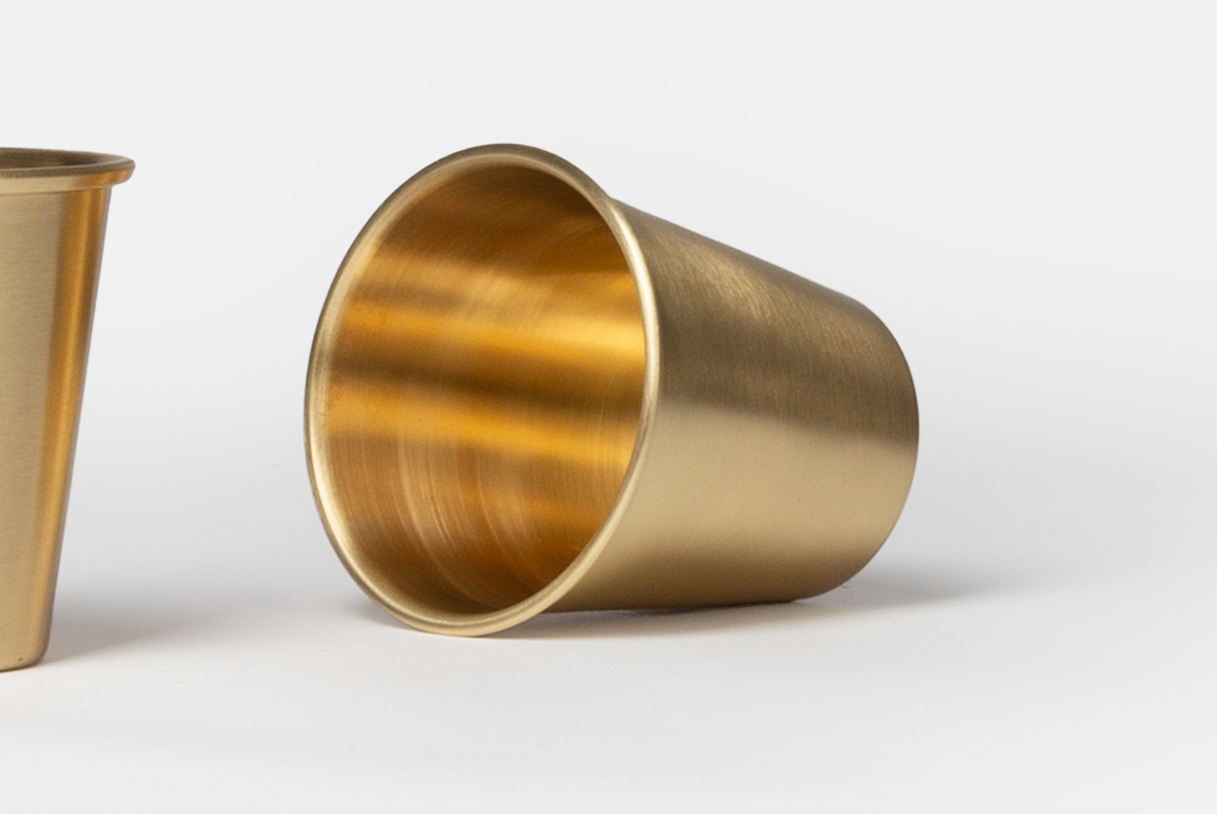 Brass Cup by Japanese Brand Fog Linen, shown as a pair with one cup standing and one laying down close up, on a white background